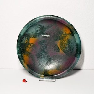 Peut inclure: Une assiette décorative ronde avec un motif tacheté dans les tons de sarcelle, de violet et d'orange. L'assiette a un bord foncé et le mot "STEELES" est visible. Une petite figurine de coccinelle est au premier plan.