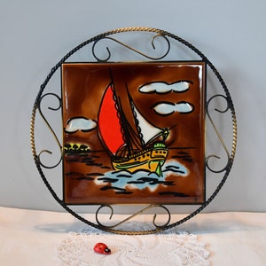 Könnte beinhalten: Eine vintage Keramikfliese mit einem gemalten Bild eines Segelboots auf dem Wasser. Die Fliese ist in einem schwarzen und goldenen Metallrahmen gefasst.