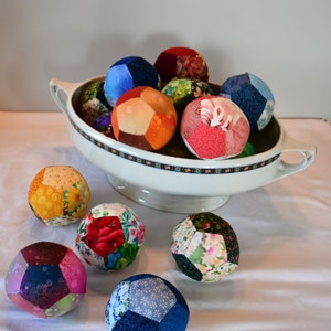 Könnte beinhalten: Eine weiße Keramikschüssel mit floralem Muster am Rand, gefüllt mit bunten Stoffkugeln. Die Kugeln bestehen aus Patchwork-Stoff in verschiedenen Farben und Mustern. Die Schüssel steht auf einer weißen Tischdecke.