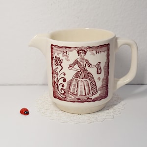 Könnte beinhalten: Cremefarbener Keramikkrug mit einer weinroten Illustration einer Frau in einem historischen Kleid. Der Krug hat einen Ausguss und einen Henkel. Ein kleiner roter Marienkäfer sitzt in der Nähe des Krugbodens.