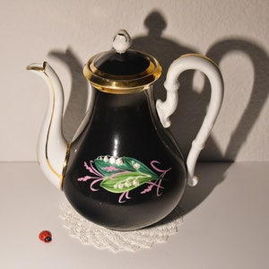 Könnte beinhalten: Eine schwarz-weiße Porzellan-Teekanne mit goldenen Verzierungen, einem weißen Griff und einem Ausguss. Die Teekanne hat ein Blumendesign mit rosa und grünen Akzenten. Eine kleine Marienkäfer-Figur befindet sich in der Nähe der Teekanne.