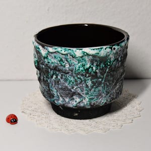 Könnte beinhalten: Ein kleiner, schwarzer Keramiktopf mit grüner und weißer Glasur. Der Topf hat eine strukturierte Oberfläche und eine abgerundete Form.