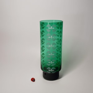 Könnte beinhalten: Ein hohes, grünes Glas mit schwarzem Sockel, verziert mit einem sich wiederholenden silbernen Blattmuster. Das zylindrische Glas steht aufrecht. Ein kleiner, roter Marienkäfer befindet sich auf der Oberfläche in der Nähe des Glases.