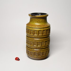 Op de afbeelding: Een cilindrische keramische vaas met een gespikkeld mosterdgeel en bruin glazuur, voorzien van drie horizontale banden met reliëf vierkante patronen met centrale stippen. Naast de vaas staat een klein, rood en zwart decoratief lieveheersbeestje.