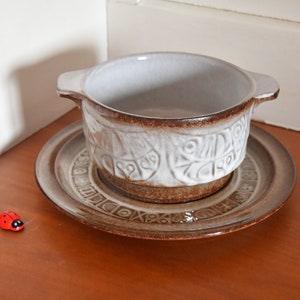 Könnte beinhalten: Eine weiße Keramikschüssel mit braunem Rand und einem geometrischen Muster. Die Schüssel steht auf einer passenden braunen und weißen Untertasse. Ein kleiner roter Marienkäfer befindet sich auf dem Tisch.