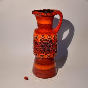 Könnte beinhalten: Eine hohe, orange-braune Keramikvase mit einem strukturierten Design. Die Vase hat eine breite Öffnung und einen schmalen Hals. Sie ist mit einem sich wiederholenden Muster aus Blumen und Blättern verziert.