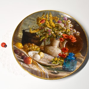 Può includere: Un piatto decorativo con un dipinto di natura morta raffigurante un bouquet di fiori, frutta e una bottiglia di vetro blu. Il piatto ha un bordo dorato ed è posto su uno sfondo bianco. Una piccola coccinella è a sinistra.