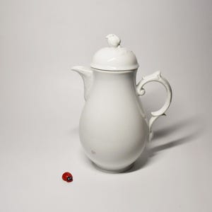 Op de afbeelding: Een witte porseleinen theepot met een glad, rond lichaam, een gebogen handvat en een deksel bekroond met een klein vogelfiguurtje. Naast de theepot staat een minuscuul rood met zwart lieveheersbeestje als figuurtje.