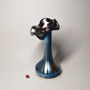 May include: A blue and silver glass vase with a ruffled top. The vase has vertical stripes and a bulbous base. A small red ladybird sits near the base of the vase. The vase is on a white doily.