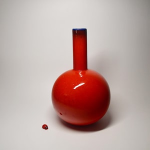 Könnte beinhalten: Eine leuchtend rote, kugelförmige Vase mit einem langen, schmalen Hals und einem dunkelblauen Rand. Neben dem Fuß der Vase befindet sich eine winzige, rote Marienkäfer-Figur mit schwarzen Punkten und einem schwarzen Kopf, die einen Maßstabsunterschied zeigt.