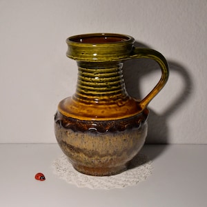 Könnte beinhalten: Eine braune und grüne Keramikvase mit einer breiten Öffnung und einem Henkel. Die Vase hat eine strukturierte Oberfläche und eine braune Glasur.