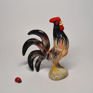 Könnte beinhalten: Eine Hahn-Figur aus Keramik mit glänzender Oberfläche, ausgestattet mit einem leuchtend roten Kamm und Kehllappen, einem schwarzen Hals sowie einem Schwanz mit schwarzen, braunen und cremefarbenen Federn. Links daneben steht ein kleiner Marienkäfer.