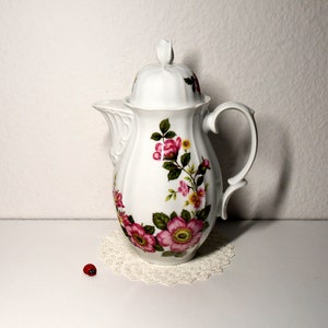 Könnte beinhalten: Weiße Porzellan-Kaffeekanne mit floralem Design mit rosa und gelben Rosen. Die Kanne hat einen Deckel und einen Henkel.