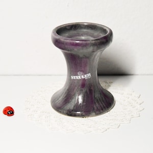 Könnte beinhalten: Ein lila-grauer Kerzenhalter aus Keramik mit ausgestelltem Boden und Oberteil. Der Kerzenhalter hat den Aufdruck "STEULER design" auf der Seite. Ein kleiner roter Marienkäfer sitzt links vom Kerzenhalter.