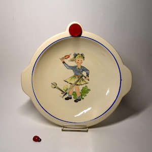 Può includere: Un piatto in ceramica bianca con un bordo blu e un'illustrazione di una ragazza che guida uno scooter. Il piatto ha una manopola rossa nella parte superiore.