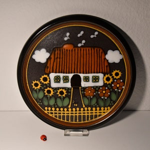 Op de afbeelding: Een ronde keramische wandplaat met een bruine achtergrond en een wit huis met een rood dak en gele ramen. Het huis is omgeven door een geel hek en groene planten met oranje en gele bloemen. Er zijn witte wolken en bruine vogels in de lucht.