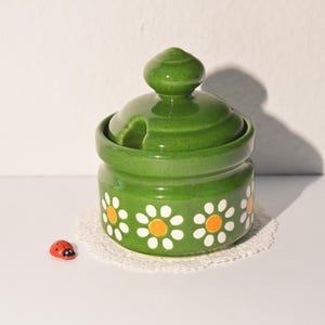 Könnte beinhalten: Ein grüner Keramiktopf mit Deckel, verziert mit weißen Gänseblümchen mit orangefarbenen Zentren. Ein kleiner roter Marienkäfer befindet sich in der Nähe. Der Topf steht auf einer weißen Spitze vor weißem Hintergrund.