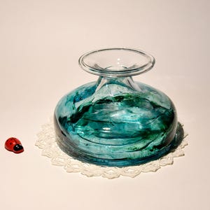 May include: A teal blue glass vase with green swirls and a wide, round base. The vase is sitting on a white crocheted doily.