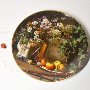 Può includere: Un piatto decorativo con una pittura di natura morta di fiori, frutta e fogliame. Il piatto ha un bordo dorato e uno sfondo scuro. La frutta comprende mele rosse e gialle. Una piccola coccinella rossa è sulla superficie bianca.