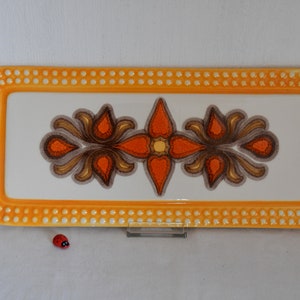 Könnte beinhalten: Ein rechteckiges Serviertablett aus Keramik mit gelbem Rand und weißem Zentrum. Das Zentrum zeigt ein stilisiertes Blumenmuster in Orange-, Braun- und Gelbtönen. Der Rand ist mit kleinen weißen Punkten verziert.