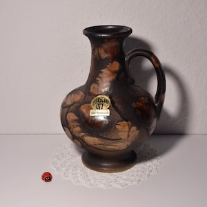 Könnte beinhalten: Eine braune Keramikvase mit einem marmorierten Überzug und einem Henkel. Die Vase hat ein Etikett mit der Aufschrift "Ruscha Art" und "echt Handarbeit".