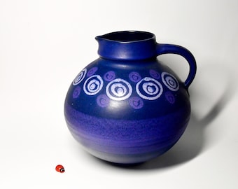 May include: A blue ceramic pitcher with a white and purple design of concentric circles. The pitcher has a round body and a handle.