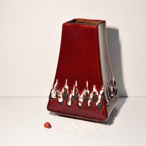 Peut inclure: Un vase en céramique bordeaux avec un dessus carré et une base évasée. Le vase présente un motif texturé blanc et marron le long du bord inférieur. Une petite figurine de coccinelle repose près du vase.