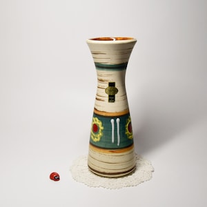 May include: A vintage ceramic vase with a beige, brown, and teal color scheme. The vase features a unique hourglass shape with a textured surface and decorative floral accents. A small sticker is affixed to the vase. A small red ladybug figurine is next to the vase.