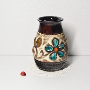 Może przedstawiać: Brązowy i beżowy ceramiczny wazon z ciemnobrązową obręczą i podstawą. Wazon zdobi duży turkusowo-pomarańczowy kwiatowy wzór z dodatkowymi akcentami kwiatowymi. Obok stoi mała czerwona figurka biedronki.