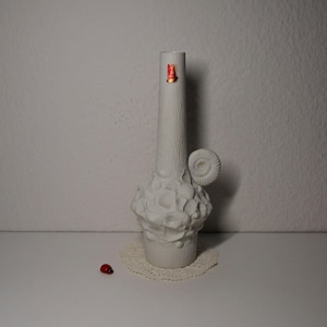 Może przedstawiać: Biały ceramiczny wazon z fakturowaną powierzchnią i wzorem przypominającym muszlę. Wazon ma wąską szyjkę i szeroką podstawę. Jest ozdobiony wypukłymi detalami przypominającymi muszle.