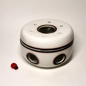 Può includere: Scaldabiberon in ceramica bianca con base rotonda e coperchio con fori. Lo scaldabiberon ha una striscia marrone intorno al centro.