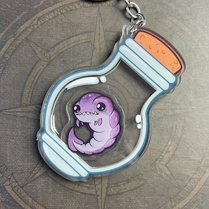 Pode incluir: Um chaveiro de plástico transparente em forma de frasco que contém uma criatura roxa e branca com olhos grandes. O frasco tem uma tampa de cortiça.