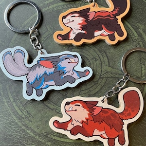 May include: Three wooden keyrings featuring cartoon animal designs. The first keyring features a red, black and white wolf running. The second keyring features a blue, grey and white wolf running. The third keyring features a red, orange and white wolf running.