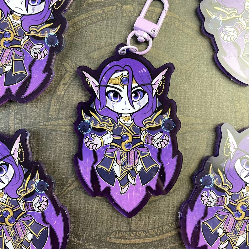 May include: Purple and gold anime character keychain with a purple and black outfit and a blue gem on their chest. The character has long purple hair and blue eyes. The keychain has a silver ring and a purple and white string.