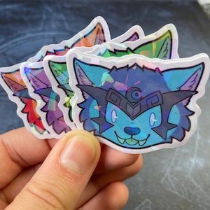 May include: Four iridescent stickers featuring a cartoon wolf with a crescent moon on its forehead. The wolf is smiling and has blue fur, a black nose, and white eyes.
