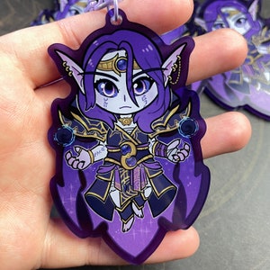 May include: Purple and gold anime character charm with a dark purple cape and a blue gem on the chest. The character has long purple hair, blue eyes, and pointed ears. The charm is made of acrylic and has a shiny finish.