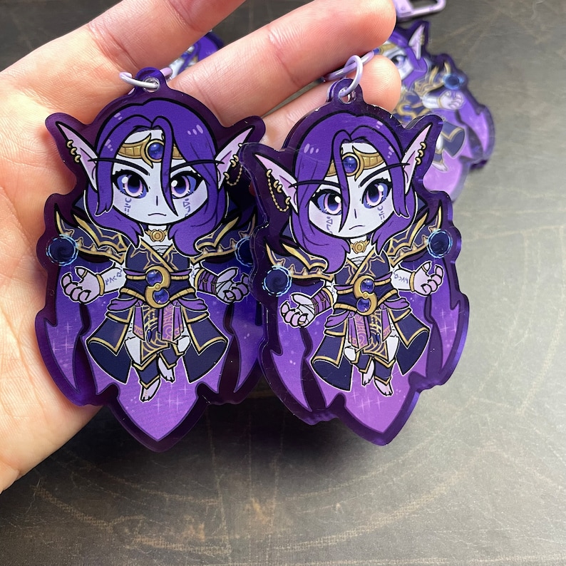 May include: Two purple and gold character charms with a sparkly background. The characters are female with long purple hair and are wearing purple and gold outfits. The charms are attached to keychains.