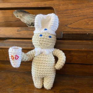 May include: A crocheted figure of a chef wearing a white hat and scarf, holding a cup with the letters "DD". The figure is beige with blue eyes and a smiling face. The background is a wooden surface.