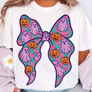 Op de afbeelding: Wit T-shirt met een groot strikdesign. De strik is versierd met een geruit patroon in teal, roze en paars, en gevuld met pompoenen en spookillustraties. Een leuke en feestelijke Halloween-thema grafiek.