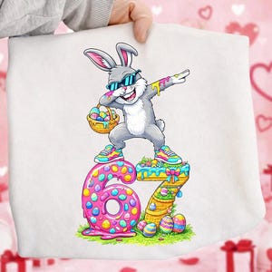 Pode incluir: Tecido branco com um gráfico colorido com tema de Páscoa. O design apresenta um coelho cinza usando óculos de sol e fazendo um dab, segurando uma cesta de ovos. O número "67" está abaixo do coelho, decorado com ovos e flores coloridas.