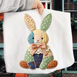 Può includere: Un coniglio patchwork colorato, realizzato con vari tessuti fantasia. Il coniglio presenta motivi floreali, a pois e a righe, un naso rosa, un fiocco a scacchi e un colletto in pizzo. È posto su uno sfondo bianco.
