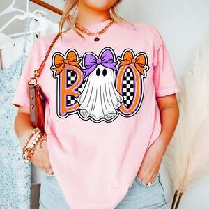 May include: A light pink t-shirt with the word "BOO" in orange and purple, and a ghost graphic. The ghost has a purple bow and is surrounded by orange bows. The letters have a checkered pattern.