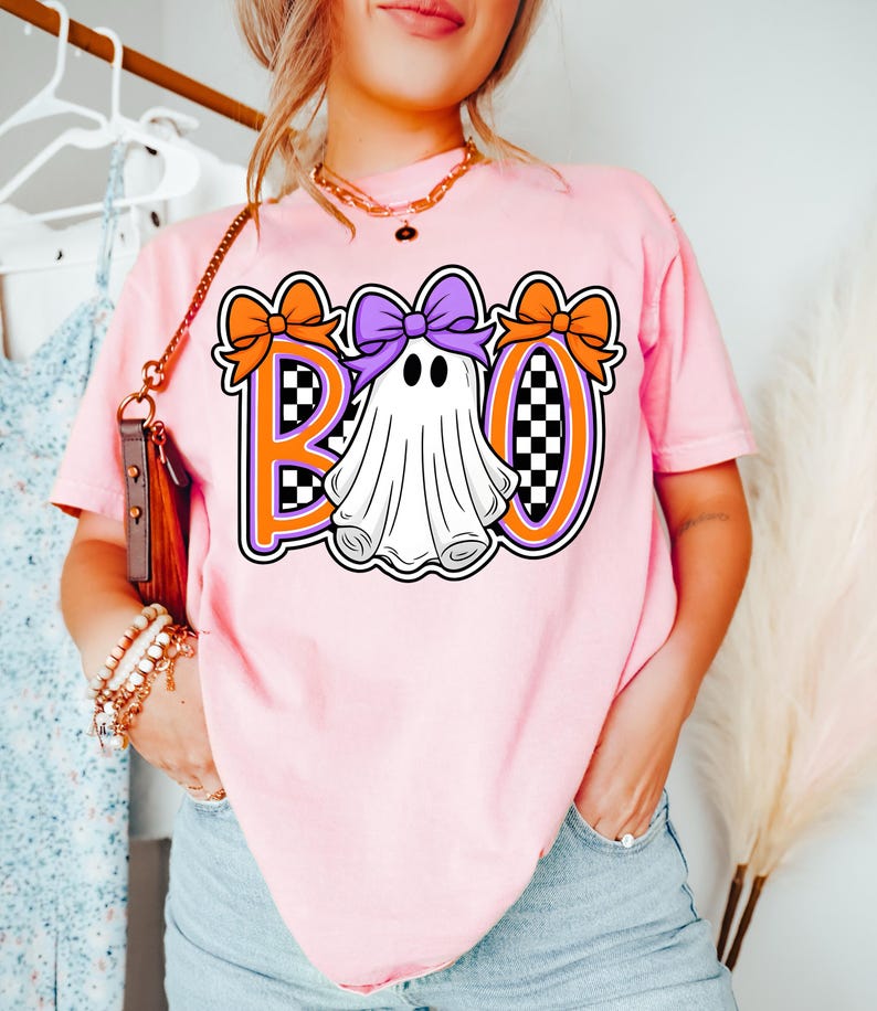 May include: A light pink t-shirt with the word "BOO" in orange and purple, and a ghost graphic. The ghost has a purple bow and is surrounded by orange bows. The letters have a checkered pattern.