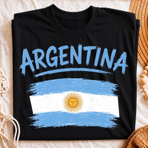 May include: A black short-sleeved t-shirt featuring the word ARGENTINA in a light blue brushstroke font above a distressed graphic of the Argentine flag, which includes horizontal light blue and white stripes and a central yellow sun emblem.