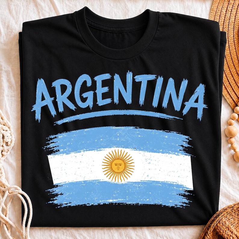 May include: A black short-sleeved t-shirt featuring the word ARGENTINA in a light blue brushstroke font above a distressed graphic of the Argentine flag, which includes horizontal light blue and white stripes and a central yellow sun emblem.