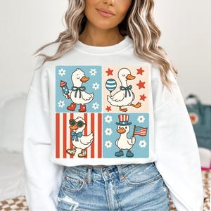 May include: White sweatshirt with a graphic of four cartoon ducks wearing patriotic accessories. The ducks are in a grid pattern with red, white, and blue stripes and stars. The text on the sweatshirt says "Duck, Duck, Goose".