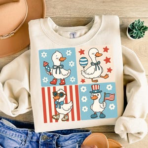 May include: A cream-colored sweatshirt with a graphic of four cartoon ducks wearing patriotic accessories. The ducks are in a grid pattern with red, white, and blue stripes and stars.