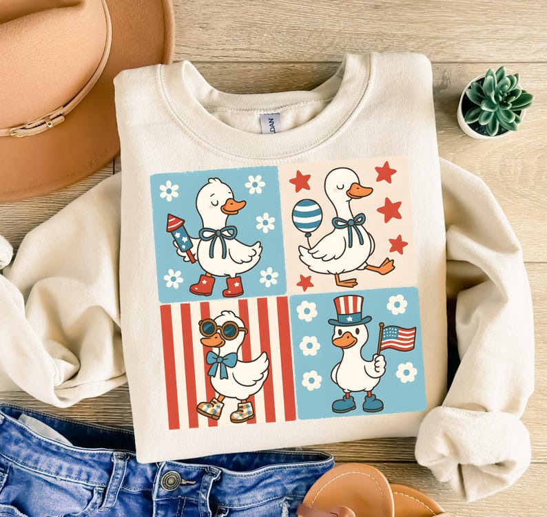 May include: A cream-colored sweatshirt with a graphic of four cartoon ducks wearing patriotic accessories. The ducks are in a grid pattern with red, white, and blue stripes and stars.