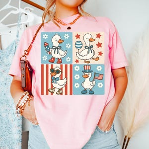 May include: Pink t-shirt with a graphic of four cartoon ducks in different patriotic outfits. The ducks are wearing sunglasses, a top hat, a bow tie, and holding a firework. The graphic is set against a red, white, and blue background.