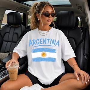 May include: A white short-sleeved t-shirt featuring the word ARGENTINA in light blue letters above a distressed graphic of the Argentine flag, which includes a central yellow sun emblem on horizontal light blue and white stripes.